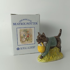 Może przedstawiać: Ceramiczna figurka psa z kolekcji Beatrix Potter. Pies jest brązowy, ubrany w zieloną kurtkę i trzyma żółty kosz. Figurka stoi na żółtej podstawie, z pudełkiem w tle.