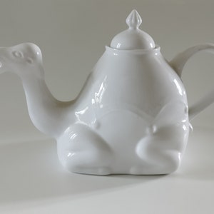 May include: A white ceramic teapot shaped like a camel. The teapot has a lid with a small knob on top and a spout that looks like the camel's head. The teapot is sitting on a white surface.