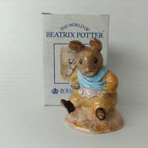 Può includere: Statuetta in ceramica di un coniglio seduto con una sciarpa blu e abiti marroni. La statuetta è su una piccola base marrone. La scatola dietro la statuetta dice "THE WORLD OF BEATRIX POTTER".