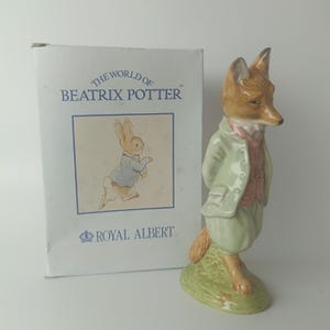 Può includere: Statuina in ceramica di una volpe con giacca e pantaloni verdi, gilet rosa e testa di volpe marrone. La statuina è su una base verde. La scatola mostra "The World of Beatrix Potter" e "Royal Albert".