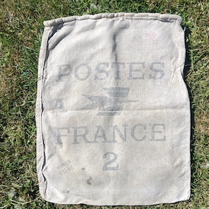 May include: A vintage beige canvas mail sack with the words "POSTES A FRANCE" printed in gray. The number "2" is printed below the text, and the bag is lying on green grass.