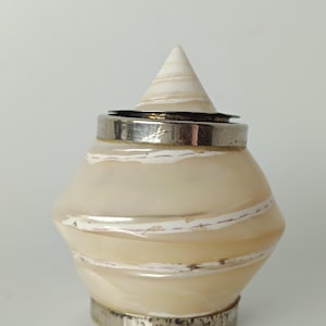 May include: A small, decorative container featuring a polished, cream-colored shell body with swirling white bands. The object is accented with a metallic silver-toned band at the base and a matching silver-toned rim, topped with a conical shell lid.