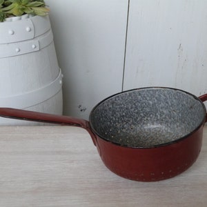 Op de afbeelding: Rode emaille zeef met een grijs gespikkeld interieur en een lange handgreep. De zeef heeft kleine gaatjes over de hele onderkant.