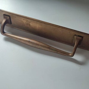 Könnte beinhalten: Ein langer, rechteckiger Türgriff aus Messing mit einem geschwungenen, glatten Griff. Der Griff ist an einer flachen Platte mit zwei sichtbaren Schrauben befestigt.