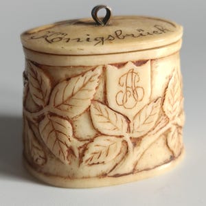 Puede incluir: Un pequeño recipiente cilíndrico blanquecino con tapa. El recipiente está tallado con un patrón de hojas y vides. La tapa tiene la palabra "Königsbrück" escrita. Un escudo con un monograma "AB" adornado también está tallado en el lateral.
