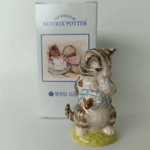 Può includere: Statuina in ceramica di un gattino della collezione "The World of Beatrix Potter". Il gattino, marrone e bianco, tiene in mano un cestino blu e bianco con un piccolo topo marrone. La statuina è su una base verde, con il logo Royal Albert.
