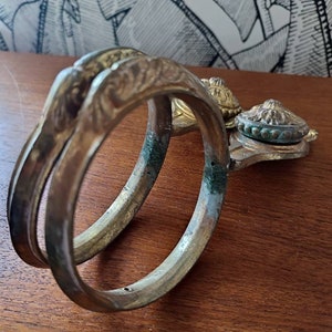 May include: A pair of antique gold-toned metal towel rings with ornate detailing. The rings are joined together by a decorative metal piece with a green and gold knob.