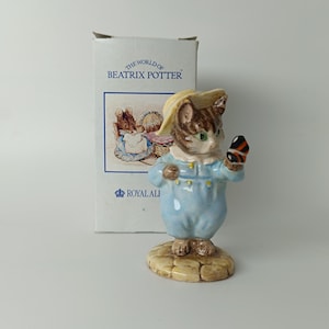 Può includere: Statuetta in ceramica di un gatto della collezione "The World of Beatrix Potter". Il gatto indossa un completo azzurro e un cappello giallo, tenendo in mano un piccolo oggetto. La statuetta è su una base strutturata, con il logo Royal Ali visibile.