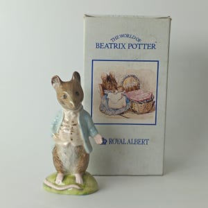 Può includere: Una statuina Royal Albert di un personaggio di topo di The World of Beatrix Potter. Il topo indossa una giacca blu e un gilet bianco, in piedi su una base verde. La scatola presenta un'illustrazione di topi e il testo "The World of Beatrix Potter".