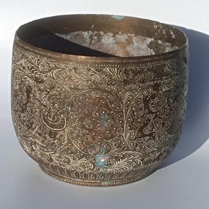 Puede incluir: Un cuenco de latón vintage con intrincados diseños florales y geométricos grabados en la superficie. El cuenco tiene una pátina ligeramente desgastada y una forma redondeada.