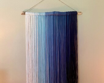 May include: A wall hanging made of yarn in shades of blue, white, and gray. The yarn is tied to a piece of driftwood, which is attached to the wall with a string.