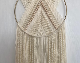 May include: A white and gold macrame wall hanging with wooden beads. The macrame is made with a variety of textures and is hung from a gold hoop.