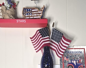 May include: Three wall-mounted shelves in red, white, and blue with the words STARS, AND, and STRIPES printed on them. The shelves hold a burlap-wrapped floral arrangement, a map cutout, two American flags, a blue vase, and a soldier figurine.