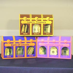 May include: Three rectangular treasure chest-shaped money boxes with three compartments labeled Save, Spend, and Give. The top box is brown and yellow with Isaac's Treasure. The bottom left is blue and orange with Bank Of Lincoln. The bottom right is purple and pink with Charlotte's Treasure.