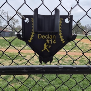 May include: A black baseball-themed holder with the name "Declan" and the number "14" in yellow. The holder features a baseball player silhouette and is attached to a chain-link fence, with a baseball field in the background.
