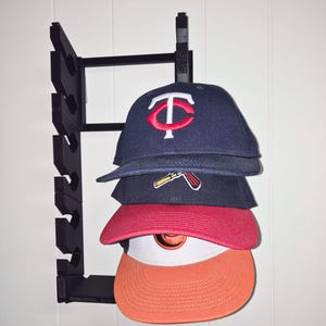 Op de afbeelding: Een zwarte aan de muur gemonteerde hoedenrek met vier baseballpetten. De petten zijn marineblauw, rood en oranje, met teamlogo's. Het rek is gemaakt van zwart metaal en is aan een witte muur bevestigd.