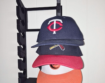 May include: A black wall-mounted hat rack holding four baseball caps. The caps are navy blue, red, and orange, with team logos. The rack is made of black metal and is mounted on a white wall.