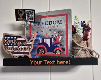 May include: A black shelf featuring a wooden cutout of the United States with the text God Bless America, a metal sign with a blue truck and the text Freedom est. 1776, a burlap-wrapped floral arrangement, and a card holder with a QR code.