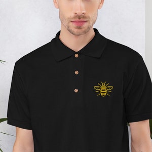 May include: Black polo shirt featuring a yellow embroidered bee motif on the left chest. The shirt has a traditional collar and three wooden buttons. The short-sleeved shirt is made of a soft material.