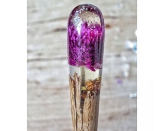 Puede incluir: Un palo de madera con una parte superior de resina transparente que contiene una flor morada prensada.