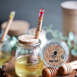 May include: Two small glass jars filled with honey, each with a cork lid and a wooden honey dipper. One jar has a cork lid with a bee design and the text "Your Text". The other jar has a wooden honey dipper with a pressed flower inside.