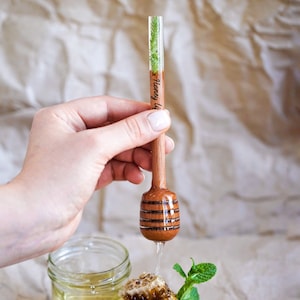 May include: A wooden honey dipper with a clear resin handle containing a small green plant. The dipper is dripping honey over a honeycomb and a jar of honey.