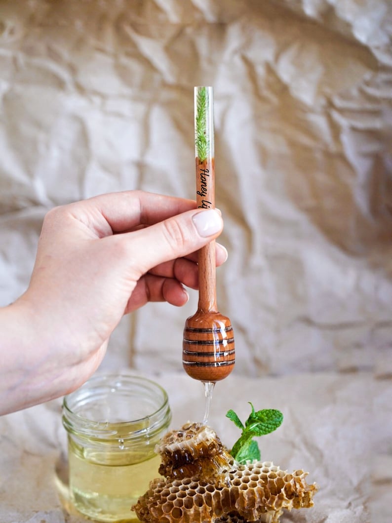 May include: A wooden honey dipper with a clear resin handle containing a small green plant. The dipper is dripping honey over a honeycomb and a jar of honey.