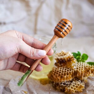 May include: A wooden honey dipper with a green resin handle, dripping honey over a honeycomb. The dipper is held in a hand.
