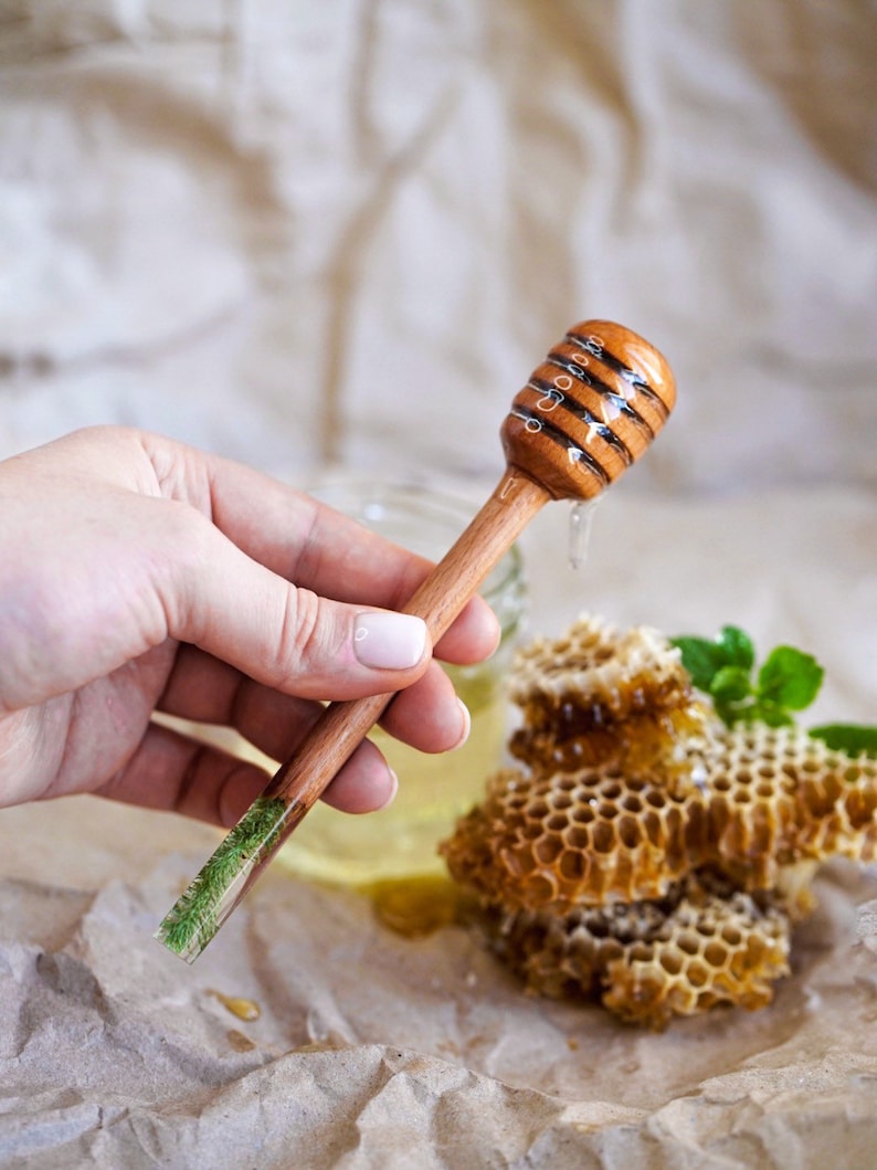 May include: A wooden honey dipper with a green resin handle, dripping honey over a honeycomb. The dipper is held in a hand.