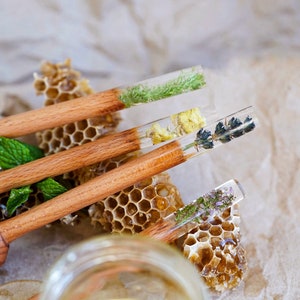 May include: Four wooden honey dippers with clear resin handles containing pressed flowers and herbs. The dippers are arranged on a honeycomb background with a jar of honey in the foreground.