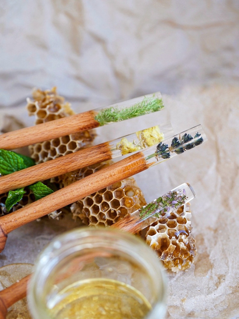 May include: Four wooden honey dippers with clear resin handles containing pressed flowers and herbs. The dippers are arranged on a honeycomb background with a jar of honey in the foreground.