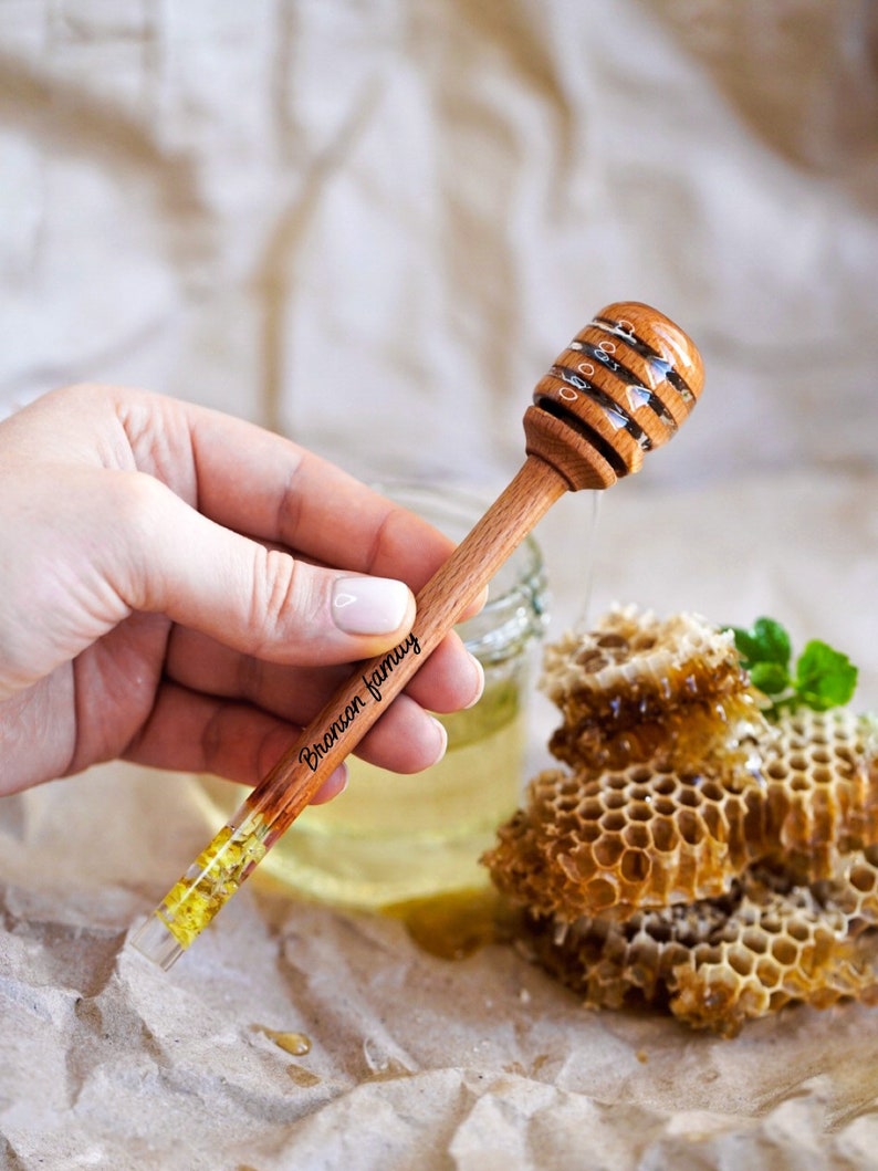 May include: A wooden honey dipper with a clear glass handle filled with yellow flower petals. The dipper is engraved with the text "Bronson family".