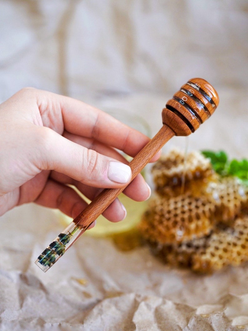 May include: A wooden honey dipper with a clear resin handle containing pressed flowers. The dipper has a traditional design with a rounded spoon-like end. The dipper is held in a hand against a background of honeycomb.