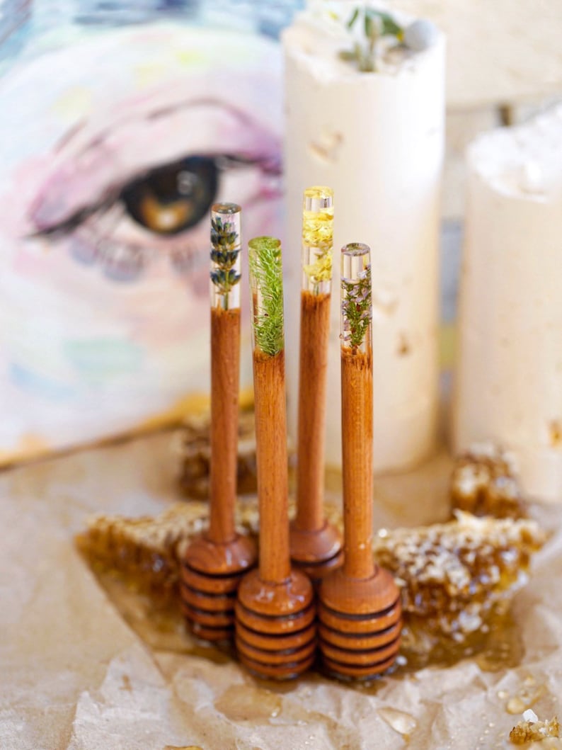 May include: Five wooden honey dippers with different pressed flowers and herbs inside the clear resin. The dippers are arranged in a row on a brown surface with honeycomb in the background.