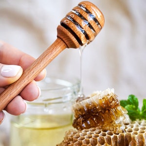 May include: A wooden honey dipper with honey dripping from it, next to a jar of honey and honeycomb.