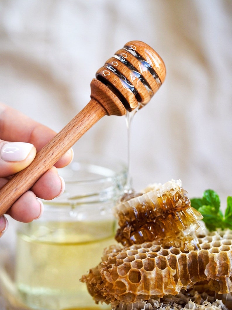 May include: A wooden honey dipper with honey dripping from it, next to a jar of honey and honeycomb.