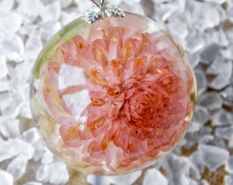 May include: A clear, spherical pendant necklace featuring a preserved pink flower encased in resin. The pendant hangs from a silver chain and is set against a background of white stones. The flower's details are visible within the transparent resin.
