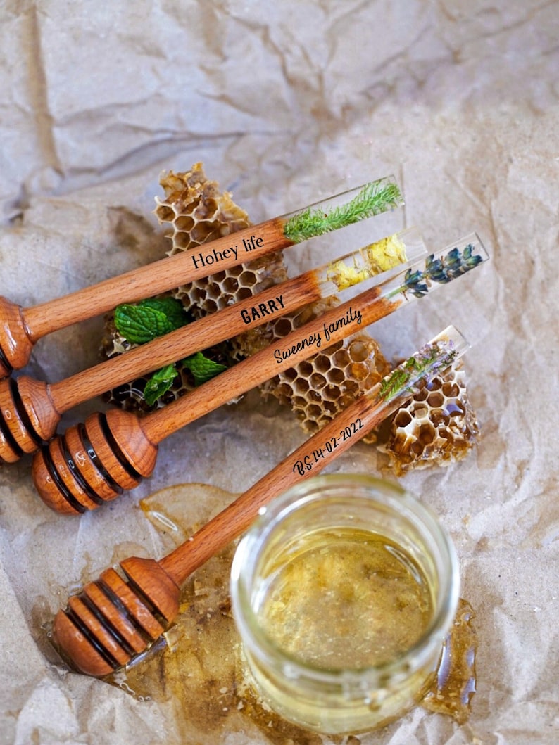 May include: Four wooden honey dippers with a clear resin handle containing pressed flowers and greenery. The dippers are labelled "Hohey life", "GARRY", "Sweeney family", and "B.S 14-02-2022". The dippers are arranged on a brown paper surface with a honeycomb and a jar of honey.