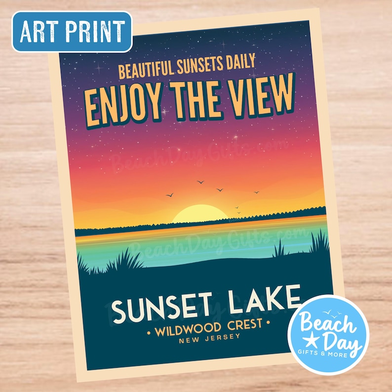 May include: Art print featuring a sunset over Sunset Lake in Wildwood Crest, New Jersey. The image displays a vibrant sky with the text "Enjoy the View" and "Beautiful Sunsets Daily". A blue circle with the text "Beach Day Gifts & More" is in the lower right corner.