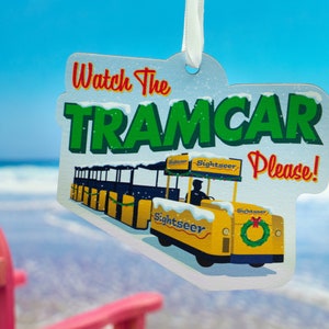Puede incluir: Un adorno de tranv&iacute;a amarillo y blanco con el texto "Watch the Tramcar Please!" sobre un fondo blanco con efecto de nieve. El tranv&iacute;a tiene la palabra "Sightseer" en el lateral.