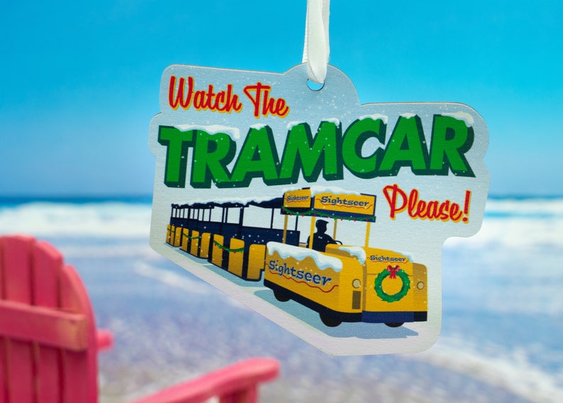 Puede incluir: Un adorno de tranv&iacute;a amarillo y blanco con el texto "Watch the Tramcar Please!" sobre un fondo blanco con efecto de nieve. El tranv&iacute;a tiene la palabra "Sightseer" en el lateral.