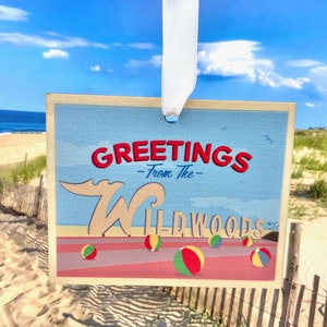 May include: A vintage-style postcard with a blue background and the text "Greetings - From The - Wildwoods" in red and white lettering. The postcard features a cartoon wave and beach balls.