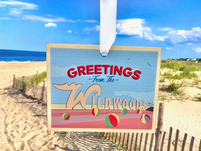 May include: A vintage-style postcard with a blue background and the text "Greetings - From The - Wildwoods" in red and white lettering. The postcard features a cartoon wave and beach balls.