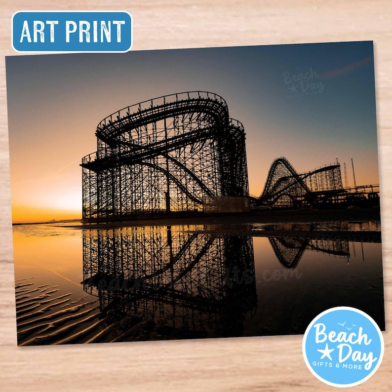May include: Art print featuring a silhouette of a wooden roller coaster at sunset, reflected in the water. The sky transitions from orange to blue. The print includes the text "ART PRINT" and a logo that says "Beach Day Gifts & More".