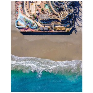 May include: Aerial view of a beach with a pier and a water park. The pier has a water slide and a roller coaster. The beach is sandy with blue water and white waves.