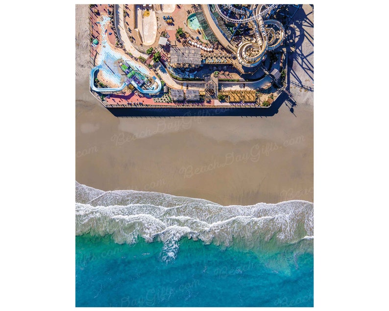 May include: Aerial view of a beach with a pier and a water park. The pier has a water slide and a roller coaster. The beach is sandy with blue water and white waves.