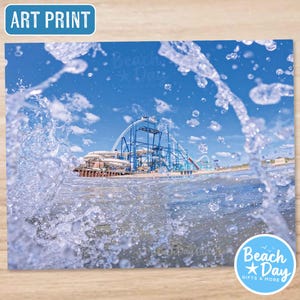 May include: Art print featuring a blue roller coaster against a bright blue sky, viewed through splashing water. The print has the text "Art Print" in a blue rectangle. The image is from Beach Day Gifts & More.