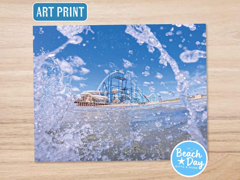 May include: Art print featuring a blue roller coaster against a bright blue sky, viewed through splashing water. The print has the text "Art Print" in a blue rectangle. The image is from Beach Day Gifts & More.