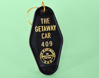 May include: A black motel-style keychain with a gold metal ring. The keychain features the text THE GETAWAY CAR 409 in gold lettering, along with a circular emblem that reads DROP IN ANY MAILBOX WE GUARANTEE POSTAGE with three small stars.