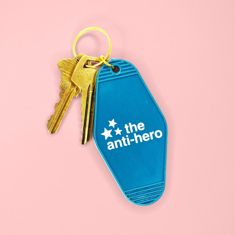Pu&ograve; includere: Un portachiavi blu a forma di chiave di motel con un anello e chiavi dorate. Il portachiavi &egrave; decorato con stelle bianche e il testo "the anti-hero" stampato su di esso.